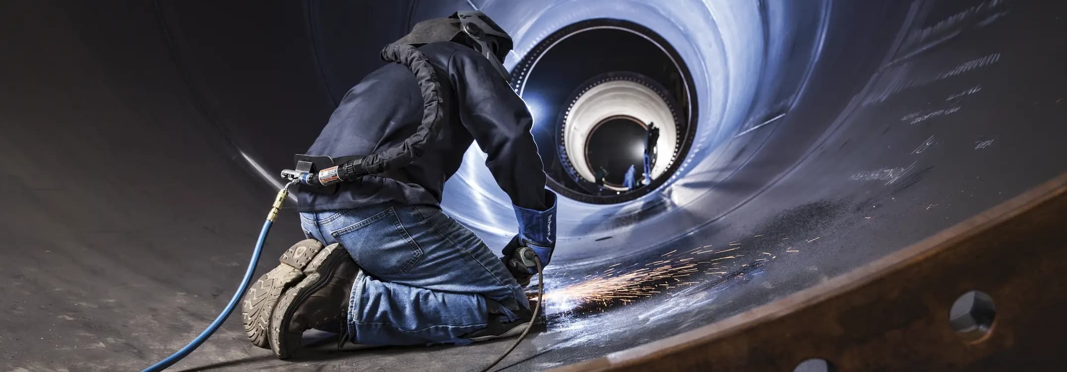 A worker performs welding inside a large cylindrical structure, surrounded by metallic walls with sparks and equipment visible.